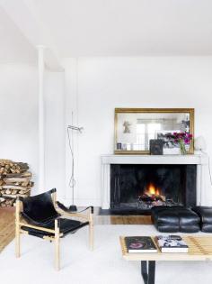 White-walled living room with black leather furniture, wooden coffee table, slink chair, fireplace, and large brass mirror on mantel.