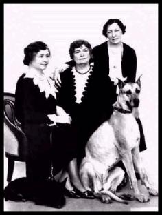This picture shows Helen Keller, Anne Sullivan Macy, and Polly Thomson, with dogs Darky and Helga, circa 1931. Helen and Anne are seated. Polly is perched on the arm of Anne's chair. Darky, a Scottish Terrier, lies curled near Helen's chair. Helga, a Great Dane, sits in front of Anne and Polly, looking toward the camera.