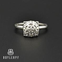 A raised solitaire setting in 14K white gold has a flower cluster of sparkling diamonds forms this distinctive Art Deco era engagement ring, circa 1930s. Elevated off the finger with a filigree type mounting, the ring is completed with a graduating band. 