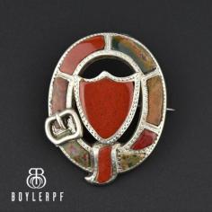 Beautiful antique Victorian Scottish agate pebble brooch with a garter buckle motif and a shield center. Colorful agates in a deep red-orange and a muted green are artfully cut and placed within the sterling silver bulbous style frame. The oval pin has an engraved border with a tubular hinge and c-clasp closure. The buckle was a symbol of the binding love between the giver and the wearer. 
