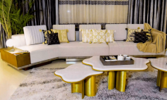 This elegant living room by MO Designs features a stylish sectional sofa with geometric and patterned throw pillows in gold, black, and cream. Gold-accented coffee tables with curved edges complement the sofa, while a soft, textured area rug and sleek decor elements complete the modern, luxurious look.

